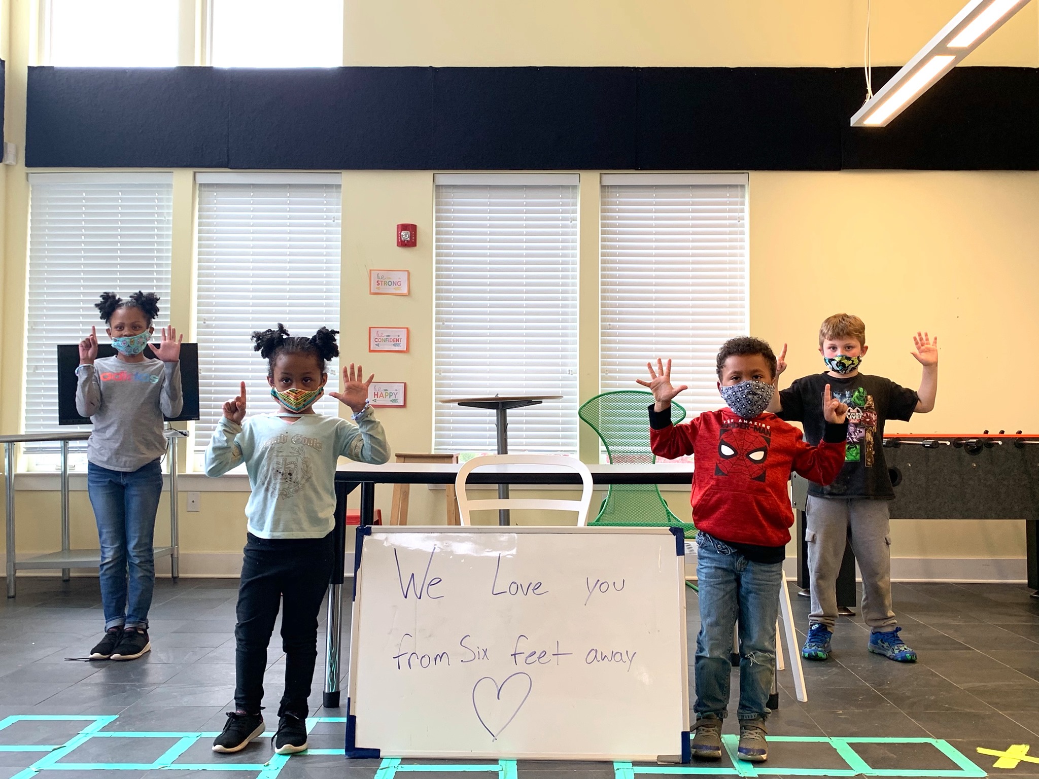 Children in masks holding six fingers up. Sign reads "we love you from six feet away."