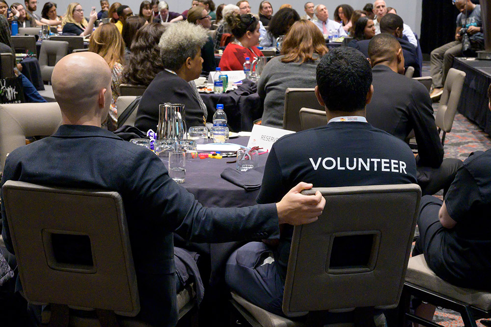 volunteers sit at a table watching a speaker on stage